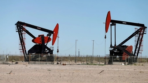 Two red and black oil pump jacks on a dry flat plain under a clear blue sky with a drilling rig visible
