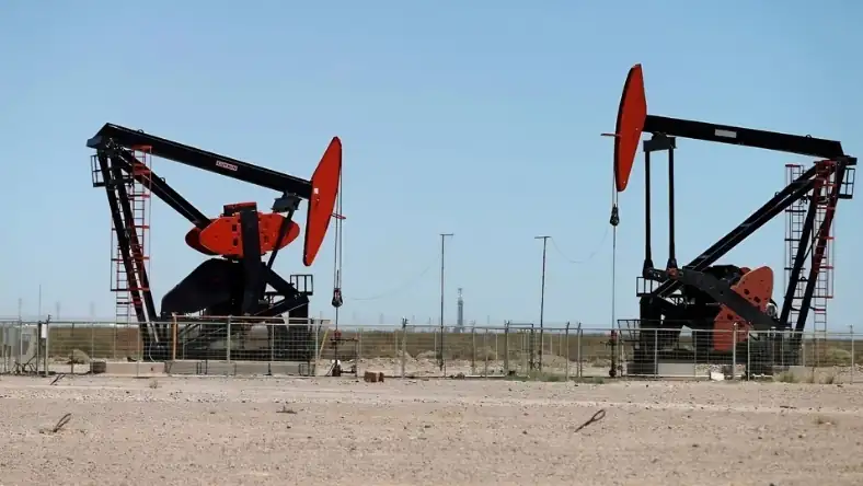Two red and black oil pump jacks on a dry flat plain under a clear blue sky with a drilling rig visible