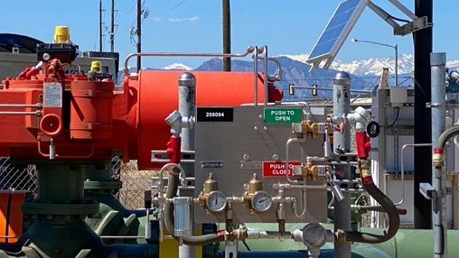 Industrial oilfield valve assembly with red vessel and pressure gauges against a clear sky and mountain range